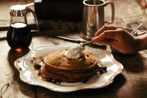 A hand adds whipped cream to a stack of pancakes on a decorative plate, with syrup and blueberries nearby.