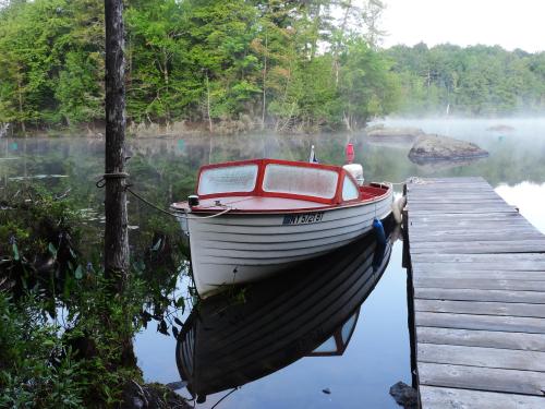 A small white boat with a red trim is docked by a wooden pier, surrounded by misty water and lush green trees.