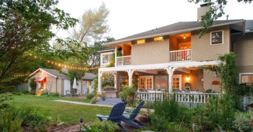 A cozy backyard scene featuring a two-story house, string lights, and a small shed surrounded by greenery.