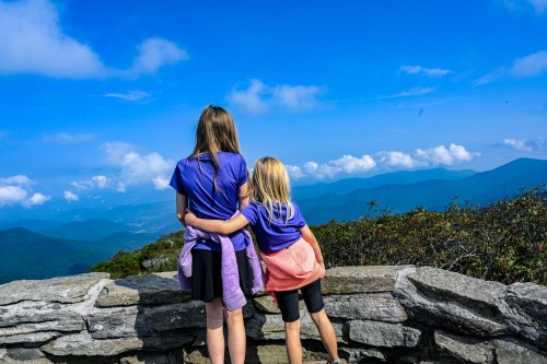 Two girls in purple shirts stand at a stone overlook, gazing at a scenic mountain view under a blue sky.
