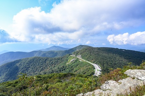 A panoramic view of lush green mountains under a partly cloudy sky, with a winding road visible in the distance.