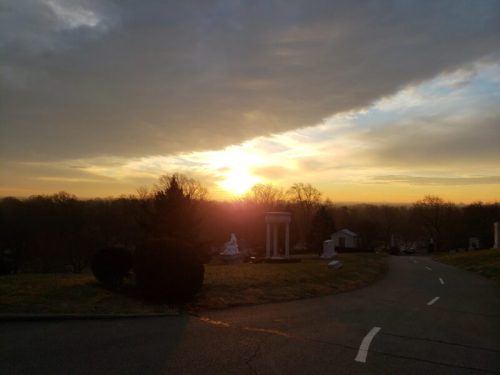 Sunset over a cemetery, with trees and a statue in the foreground, casting a warm glow against a cloudy sky.