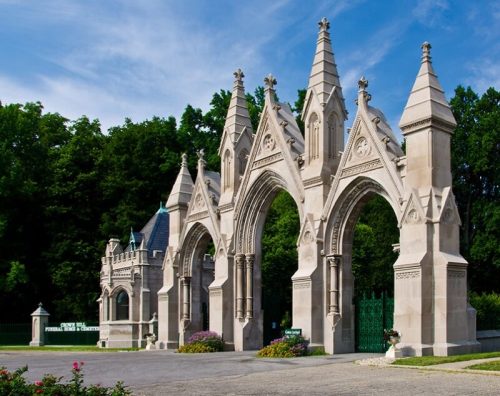 Gothic-style stone archway with intricate details, surrounded by greenery and a clear blue sky.