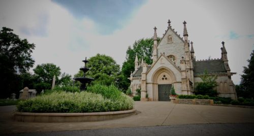 A historic stone chapel surrounded by greenery and a fountain, set against a cloudy sky.