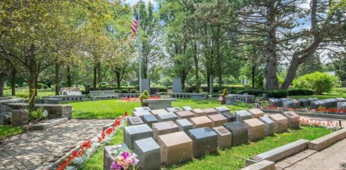 A serene cemetery scene with gravestones, flowers, and trees, featuring an American flag in the background.