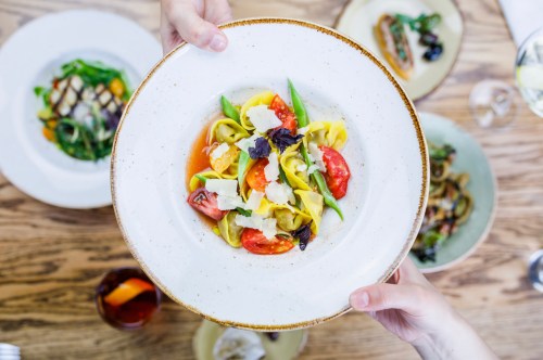 A person holds a plate of colorful pasta with tomatoes, herbs, and cheese, surrounded by other dishes on a wooden table.