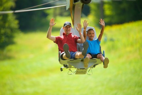 Two people, a man and a boy, smile and wave while riding a chairlift over a green landscape.