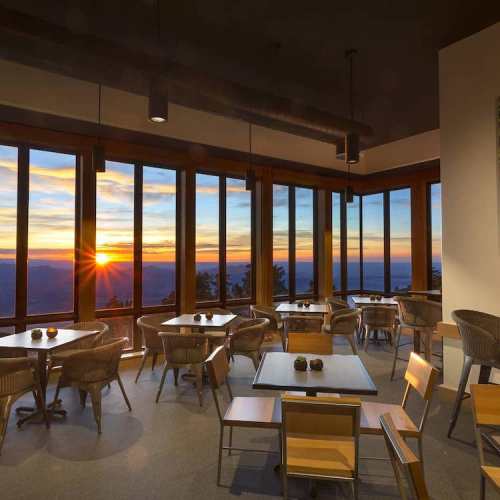 A cozy dining area with wooden tables and chairs, featuring large windows showcasing a sunset view.