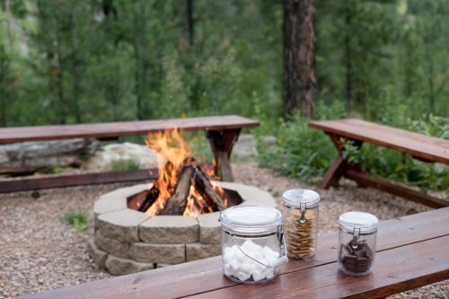 A cozy campfire surrounded by picnic tables, with jars of marshmallows, graham crackers, and chocolate nearby.