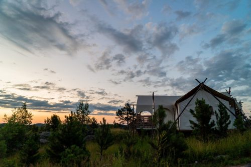 A glamping tent surrounded by greenery under a colorful sunset sky with scattered clouds.
