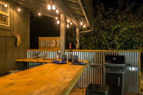 Cozy outdoor kitchen at night with string lights, wooden bar, grill, and plates set on a rustic countertop.