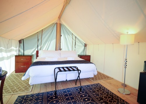 Cozy glamping tent interior with a double bed, nightstands, lamp, and decorative rugs on wooden flooring.