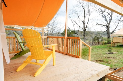 A yellow Adirondack chair on a wooden deck with a view of trees and a grassy area under an orange canopy.