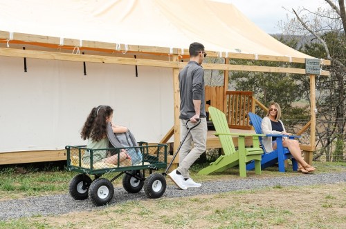 A man pulls a wagon with a child while a woman sits on a colorful chair outside a tent in a grassy area.