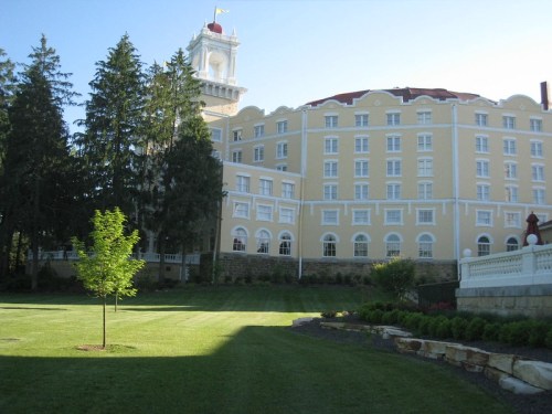 A large, historic hotel with a clock tower, surrounded by green lawns and trees under a clear blue sky.