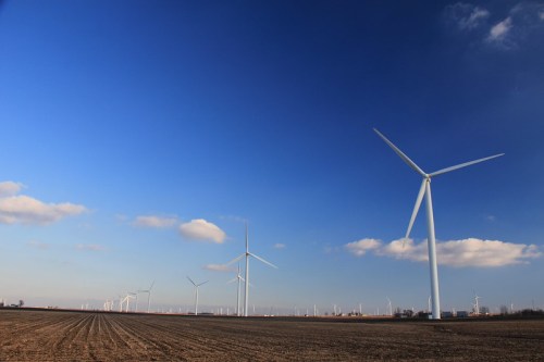 A landscape featuring several wind turbines against a clear blue sky with scattered clouds and a plowed field in the foreground.