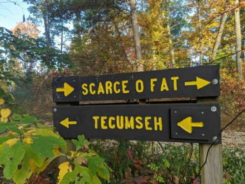 Two wooden directional signs in a forest, one pointing left to "Scarce O Fat" and the other left to "Tecumseh."