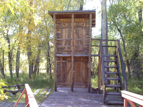 A rustic wooden restroom structure with separate entrances for women and men, surrounded by trees and greenery.