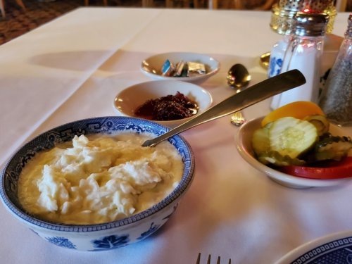 A bowl of creamy porridge with a spoon, accompanied by small dishes of jam and pickled vegetables on a table.