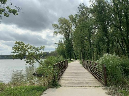 A wooden bridge leads over a calm river, surrounded by lush greenery and cloudy skies.