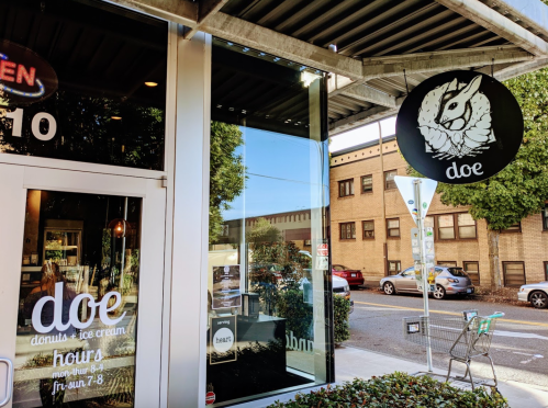Exterior of "Doe" shop featuring a sign, large windows, and a view of the street with parked cars.