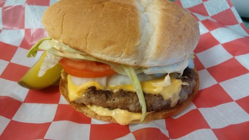 A juicy cheeseburger with lettuce, tomato, and onion on a bun, served with a pickle on a red and white checkered paper.