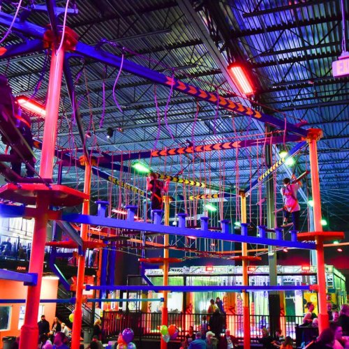 A vibrant indoor ropes course with colorful lights, featuring two people navigating the obstacles above a lively crowd below.
