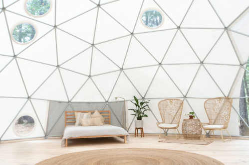 Interior of a geodesic dome with a bed, two chairs, a plant, and a round rug on a wooden floor. Natural light filters in.