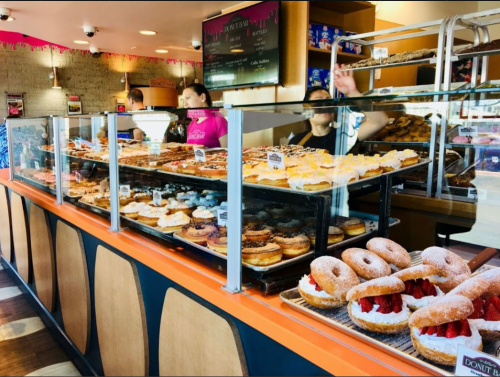 A display case filled with various donuts, with staff and customers in a bright, inviting donut shop.