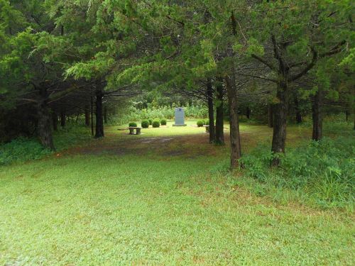 A serene clearing in a forest with a stone monument and a bench surrounded by trees and greenery.