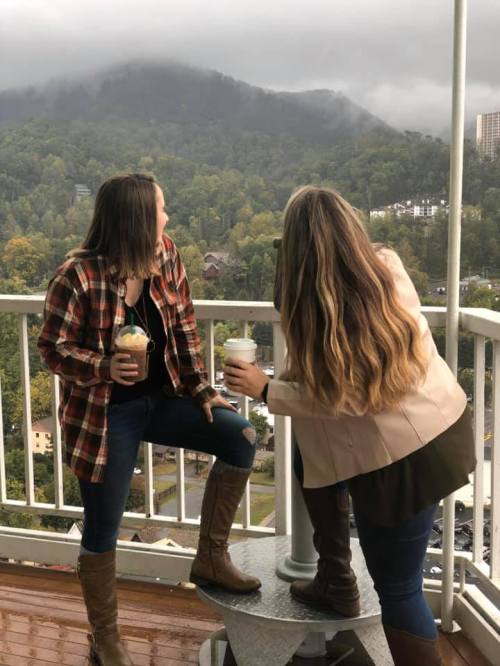 Two women on a balcony, holding drinks, looking at a misty mountain view with trees in the background.
