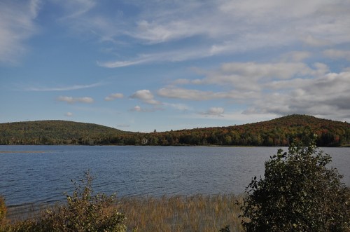 Scenic view of a calm lake surrounded by hills and colorful autumn foliage under a partly cloudy sky.