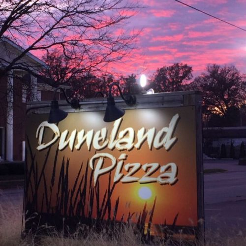Sign for Duneland Pizza with a sunset backdrop featuring vibrant pink and orange hues.
