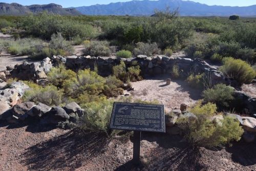 A stone circle surrounded by shrubs, with a plaque in the foreground and mountains in the background.