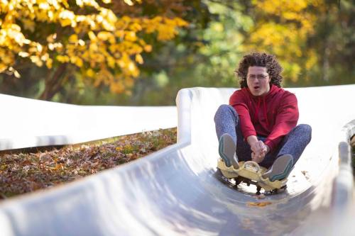 A person in a red hoodie slides down a smooth track surrounded by autumn foliage, enjoying a thrilling ride.