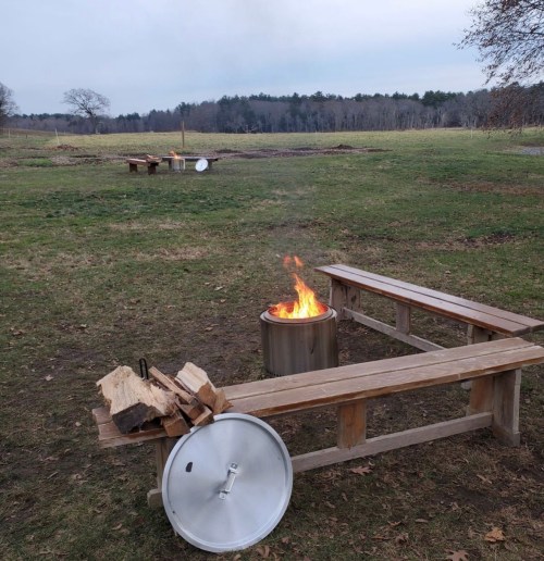 A fire burns in a metal container surrounded by wooden benches in a grassy outdoor area.