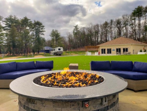 A cozy outdoor seating area with a fire pit surrounded by blue couches, set against a backdrop of trees and a building.