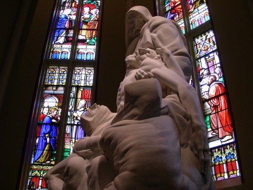 A white sculpture of a figure holding a child, set against colorful stained glass windows in a church.