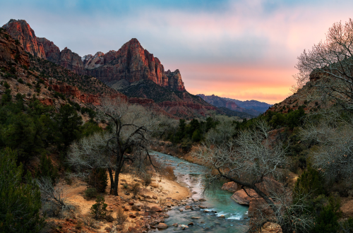 A scenic view of Zion National Park at sunset, featuring red rock formations and a winding river surrounded by trees.
