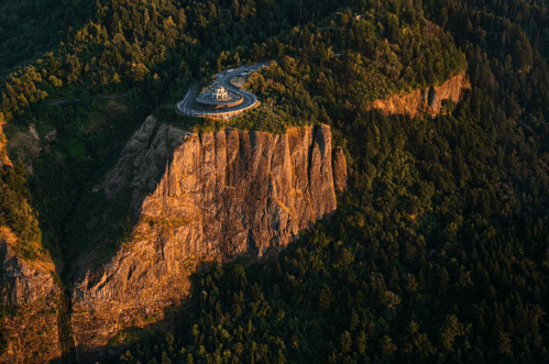 Aerial view of a winding road atop a rocky cliff surrounded by lush green trees at sunset.