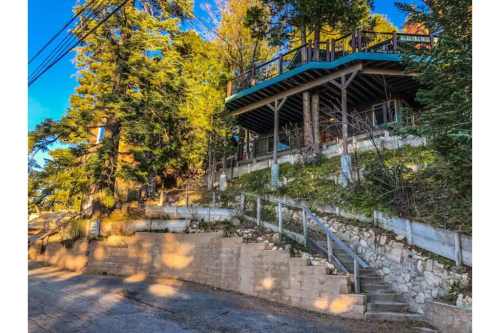 A hillside house surrounded by trees, featuring a deck and stone steps leading up from the road.