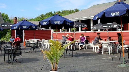 Outdoor dining area with tables and umbrellas, people enjoying meals, and a backdrop of a red building and greenery.
