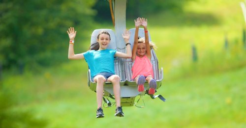 Two girls happily riding a chairlift, waving their hands, with a green landscape in the background.