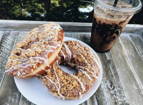 Two donuts with icing and crumb topping on a plate, next to a glass of iced coffee on a wooden table.