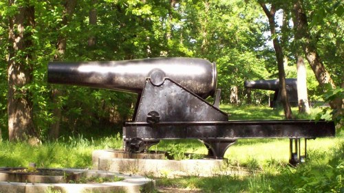 Two historic cannons positioned on stone bases, surrounded by lush green trees in a wooded area.