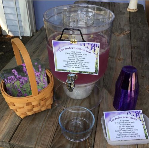 A lemonade dispenser with lavender lemonade, a basket, cups, and a recipe card on a wooden table.