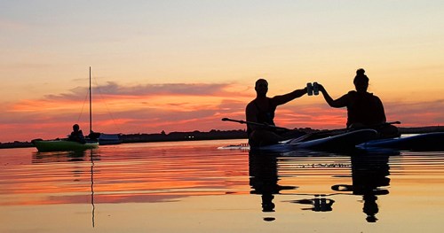 Two people in kayaks toast with drinks at sunset, with a sailboat in the background and calm water reflecting the colors.