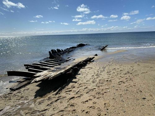 A weathered shipwreck partially submerged on a sandy beach, with calm waters and a clear blue sky in the background.