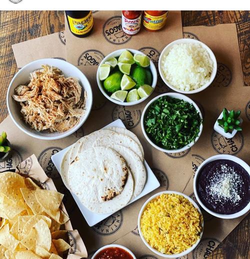 A spread of ingredients for a meal, including tortillas, shredded chicken, rice, beans, lime, cilantro, and chips.