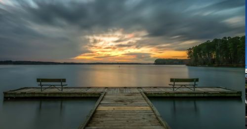 A serene lake view at sunset, featuring a wooden dock with two benches and a cloudy sky reflecting on the water.